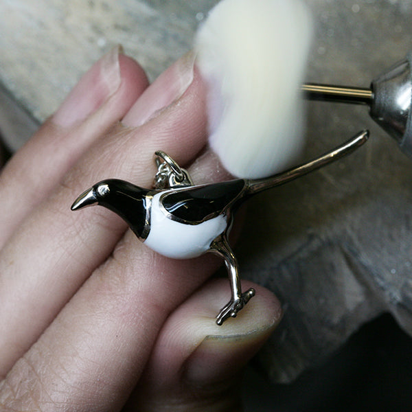 A Magpie charm with white and black enamel is being polished in the workshop with a white brush.